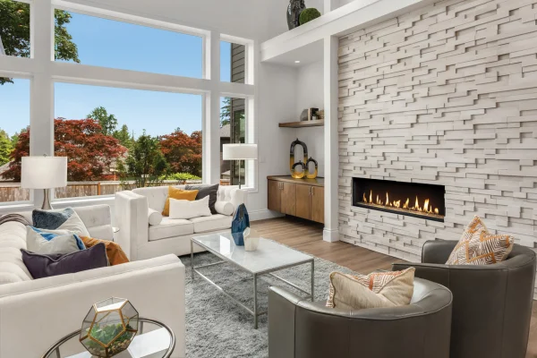Bright modern living room with large windows, white sofas, a rectangular coffee table, and a gas fireplace against a textured white stone wall.