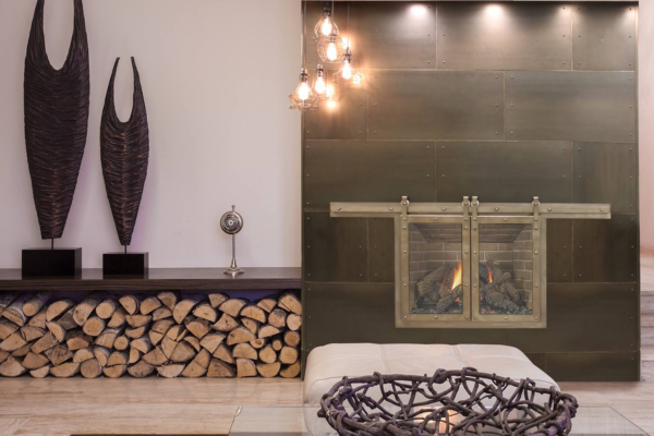 Contemporary living room with a glass-front fireplace set in dark metal panels, a stack of firewood beneath a shelf, and decorative sculptures on the mantel. A woven centerpiece sits in front of a white sofa.
