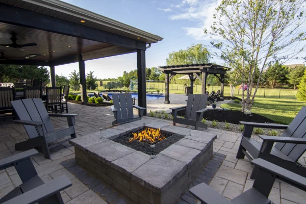 7 - Fireplace Gallery Outdoor stone patio with a square fire pit surrounded by gray Adirondack chairs; a covered seating area on the left and a wooden pergola in a grassy backyard.